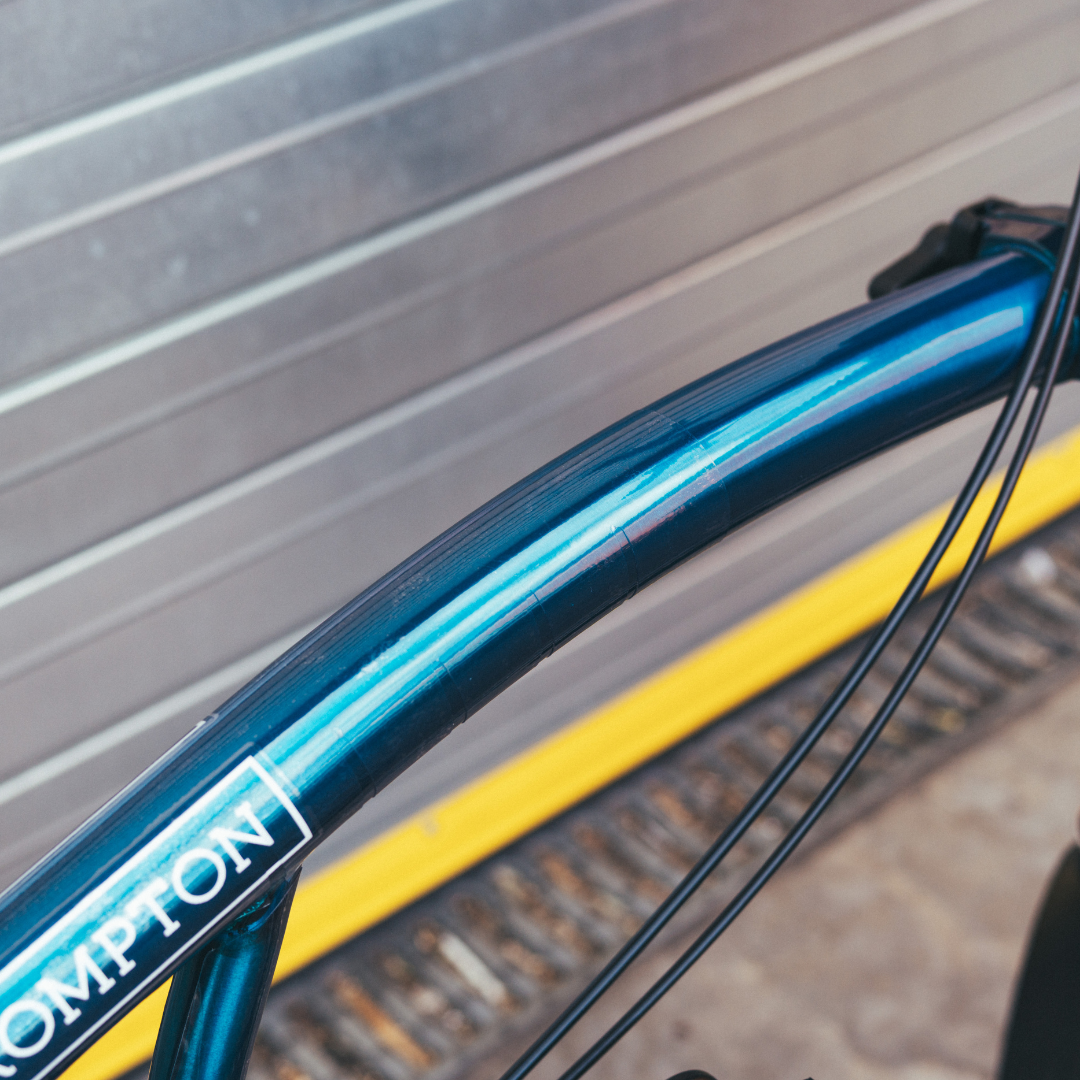 Close-up of a blue bicycle frame with 'Brompton' branding against a metallic background.