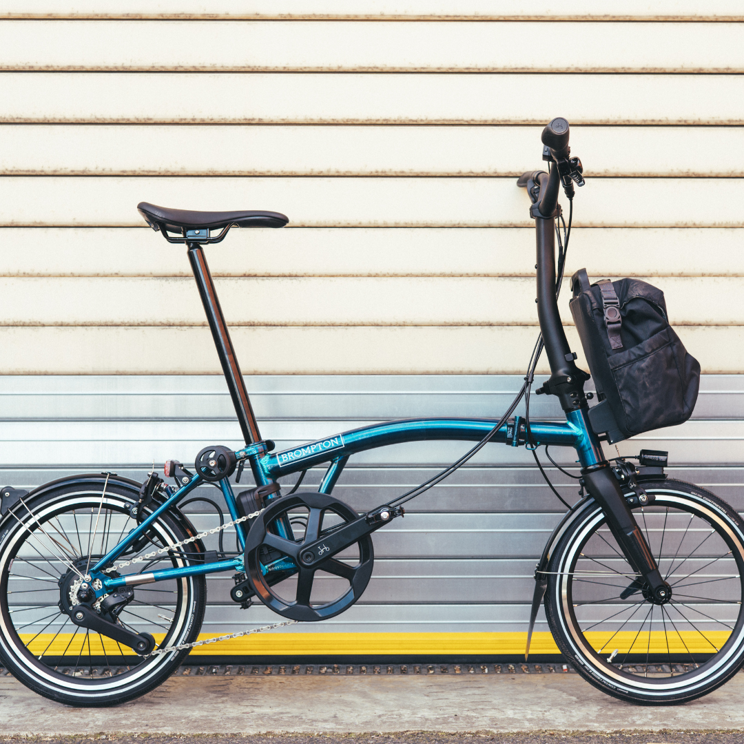 Blue folding brompton bicycle with a black bag against a metallic background