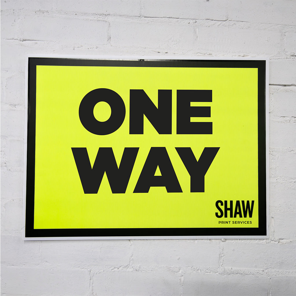 Framed poster with 'ONE WAY' text on a yellow background, mounted on a white brick wall.