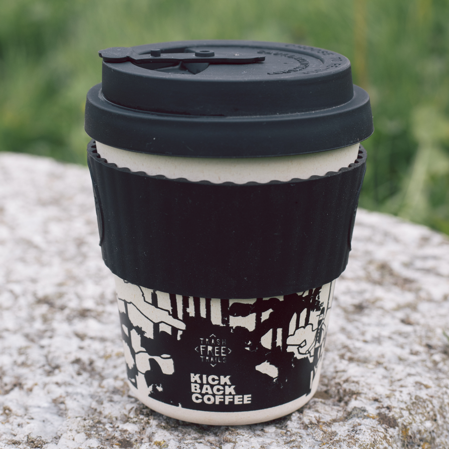 Reusable coffee cup with black lid and 'Kick Back Coffee' branding on a stone surface with grass in the background.