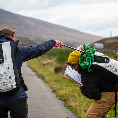 Two people outdoors on a path using a long-handled tool attached to backpacks. One person wears a navy jacket and black pants with a white backpack, while the other wears a black jacket, tan pants, and a yellow beanie with a green backpack.