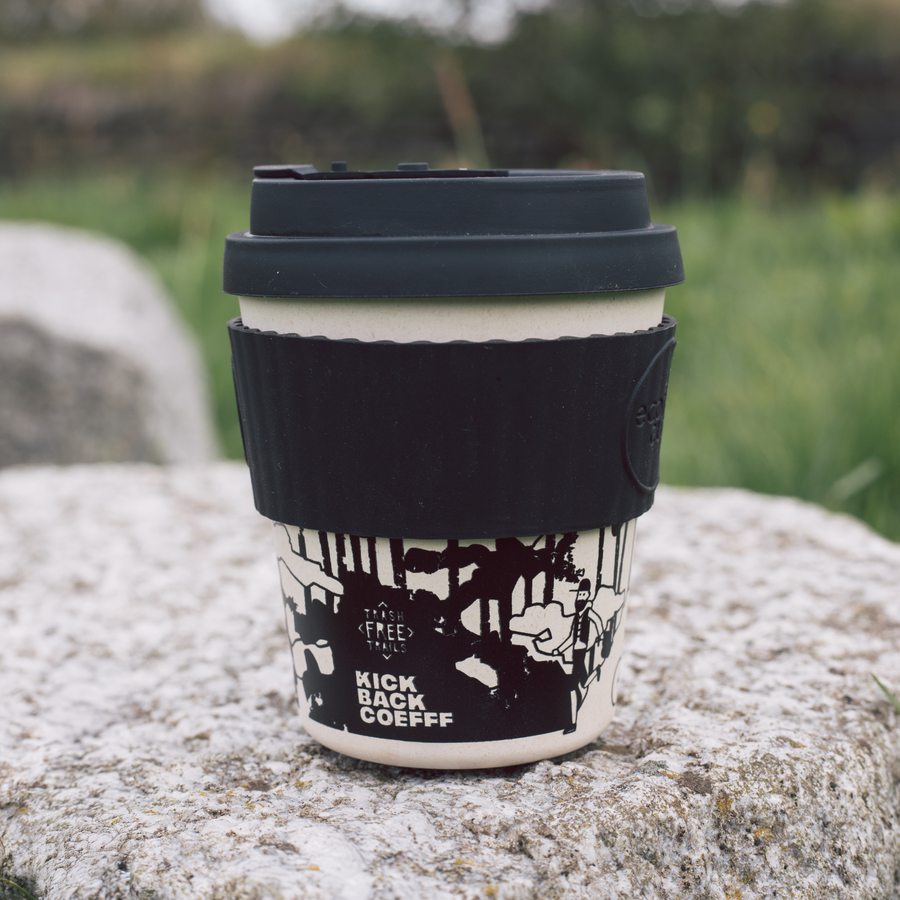 Reusable coffee cup with black lid and 'Kick Back Coffee' branding on a stone surface with grass in the background.
