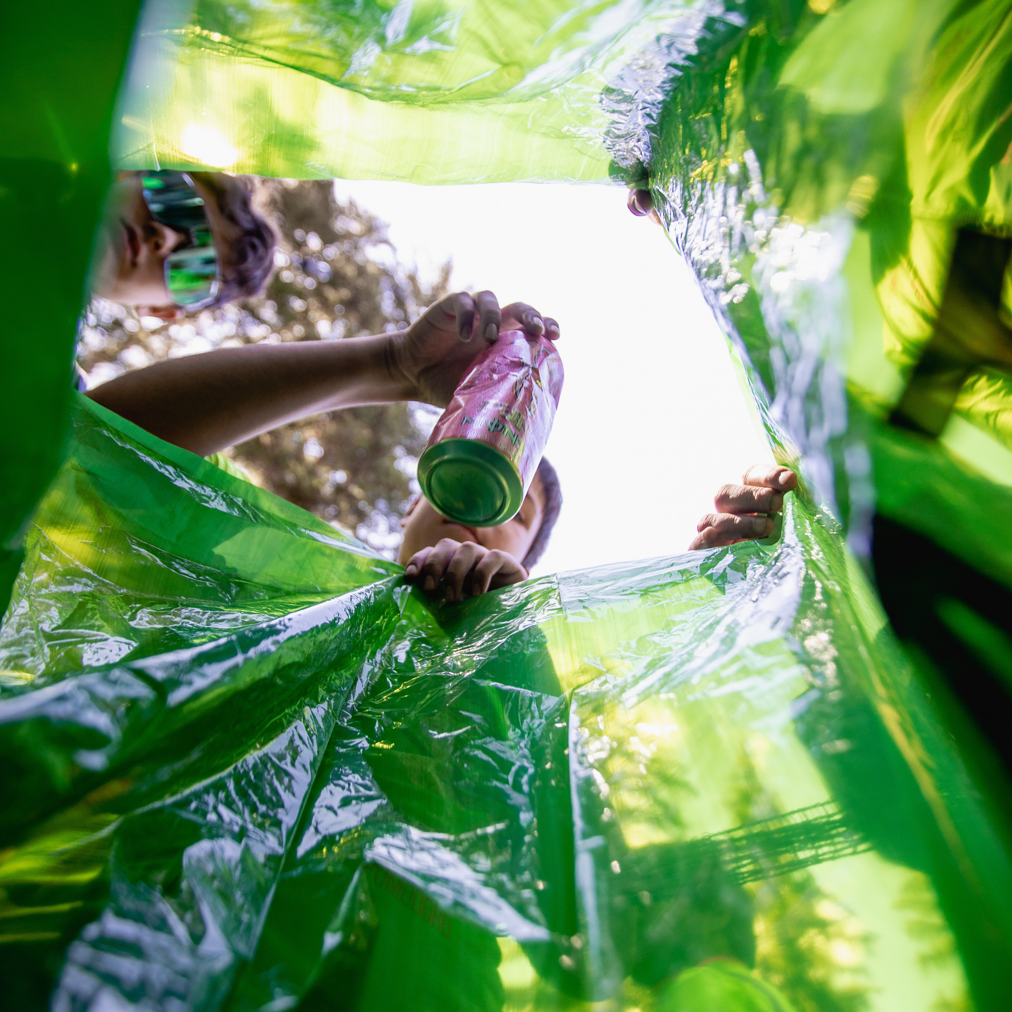 A person inside a translucent green plastic rain poncho or canopy, holding a pink beverage can up through the glossy plastic surface. The bright outdoor sky and blurred trees are visible through the transparent green material.