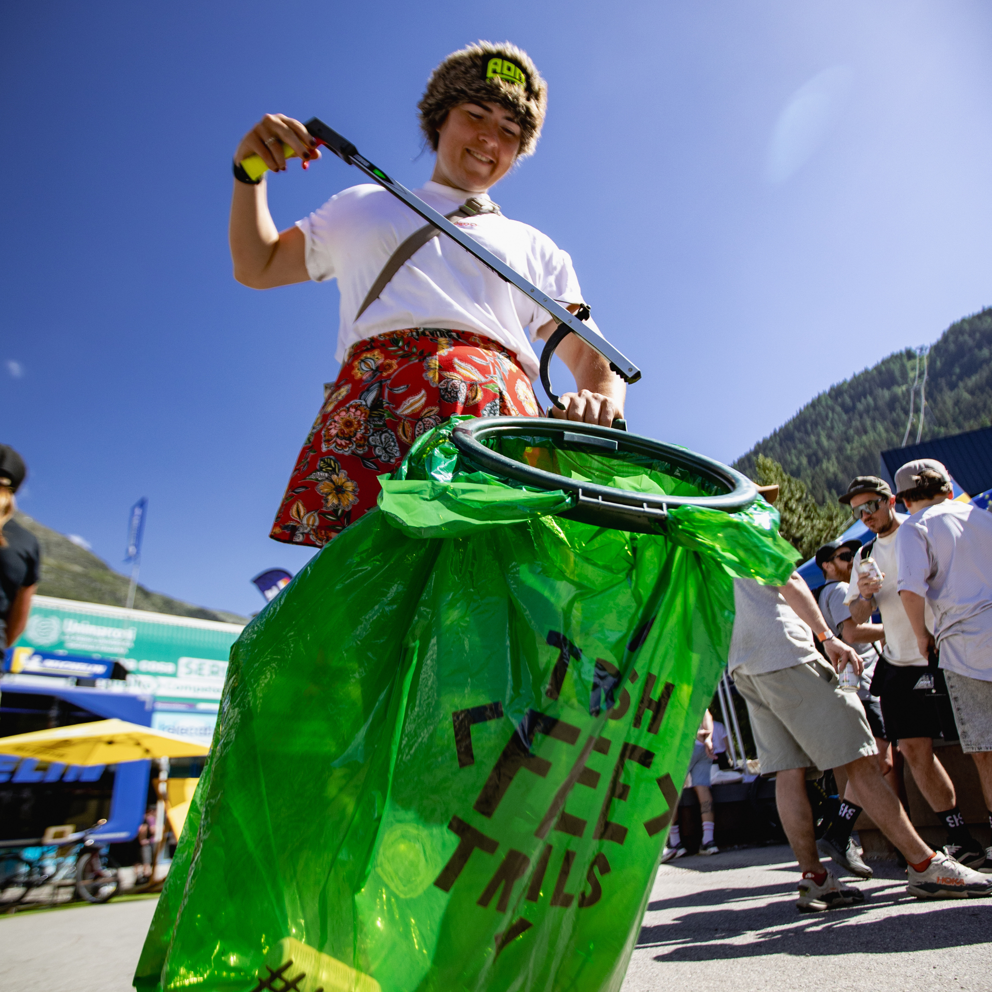 A person outdoors wearing a brown fur hat and a white shirt, holding a long-handled green feeder bag filled with bright green plastic bags. The bag has black text reading 'TSH FEED THIS'.