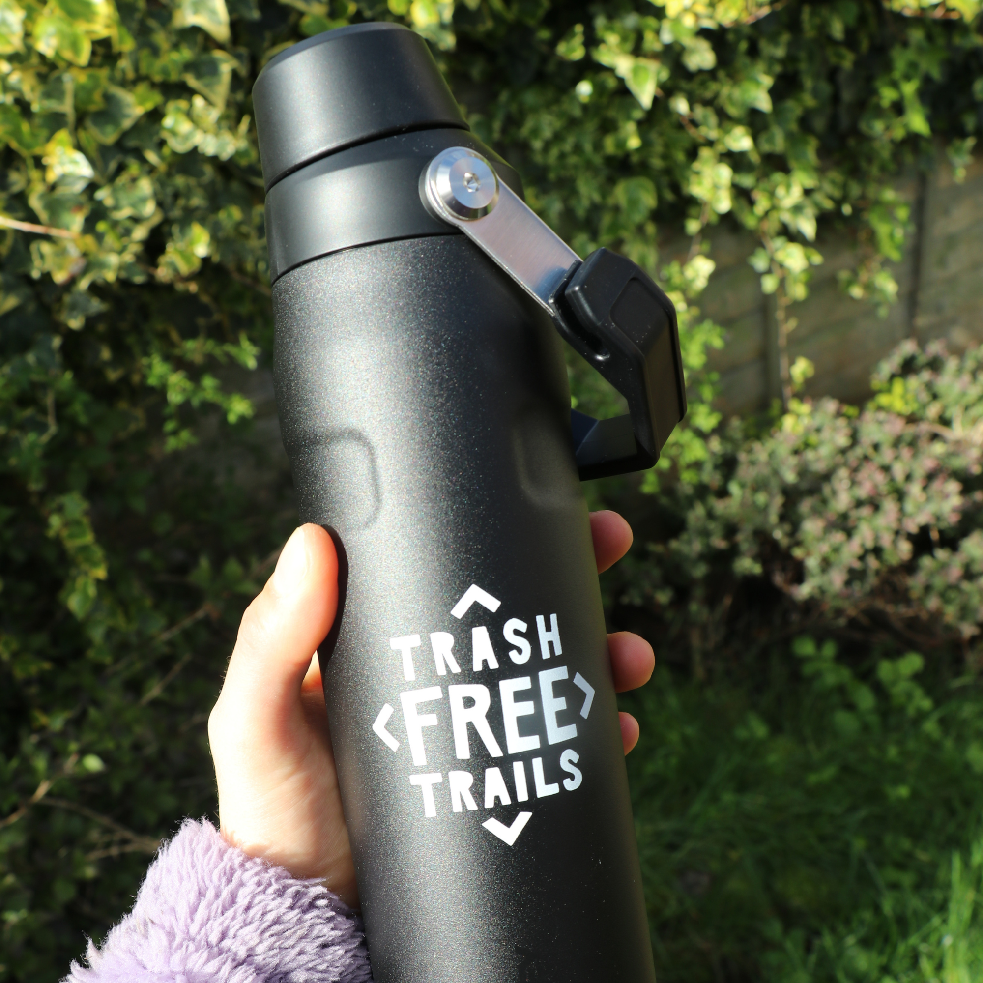 Black water bottle with 'Trash Free Trails' logo held in a hand against a green outdoor background