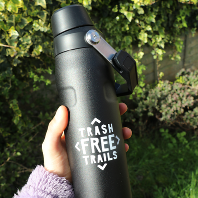 Black water bottle with 'Trash Free Trails' logo held in a hand against a green outdoor background