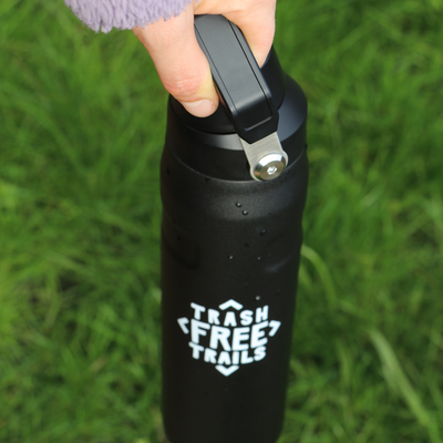 Black water bottle with 'Trash Free Trails' logo held by a hand on grass